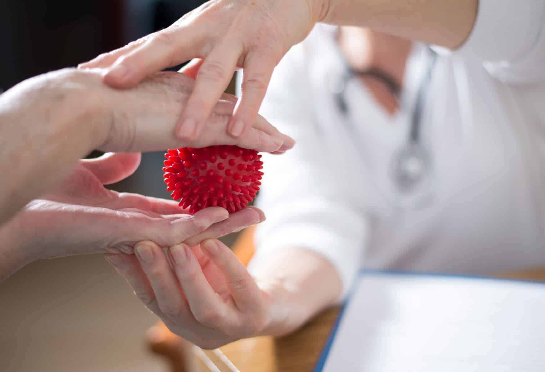 Physiotherapie bei myPhysio Deutschland: Hände einer Patientin werden von einem Physiotherapeuten bei einer Übung mit einem roten Massageball unterstützt.